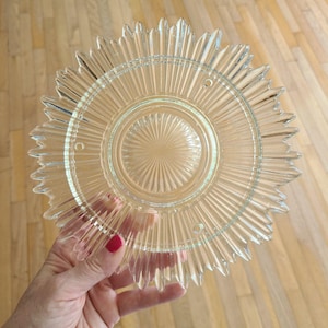 May include: A clear glass plate featuring a sunburst pattern with radiating ridges extending from a central circular medallion to a jagged, star-shaped outer edge. The design includes three small, evenly spaced circular holes near the center.