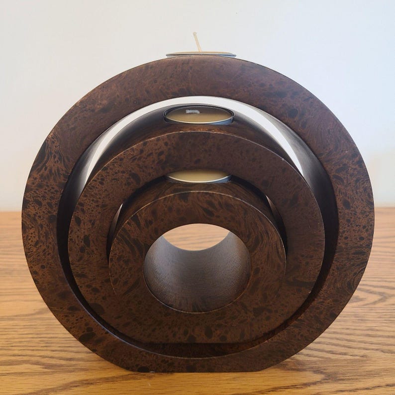 Op de afbeelding: Een houten kandelaar in donkerbruin met een uniek concentrisch cirkelontwerp. De houder heeft drie cirkelvormige niveaus, elk met ruimte voor een waxinelichtje. Bovenop staat een enkele brandende kaars.