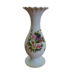 May include: A white ceramic vase with a ruffled top and a round base. The vase is adorned with a cluster of colorful, three-dimensional flowers in shades of pink, purple, yellow, and orange, with green leaves. The vase is approximately 10 inches tall.