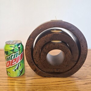 Op de afbeelding: Een donkerbruine houten kandelaar met een textuurpatroon, met drie concentrische cirkelvormige niveaus. Drie waxinelichtjes zijn in de bovenste laag geplaatst. Een groen en wit frisdrankblikje staat naast de kandelaar.