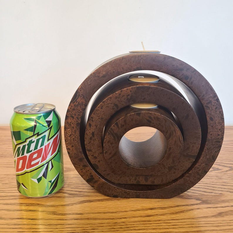 Op de afbeelding: Een donkerbruine houten kandelaar met een textuurpatroon, met drie concentrische cirkelvormige niveaus. Drie waxinelichtjes zijn in de bovenste laag geplaatst. Een groen en wit frisdrankblikje staat naast de kandelaar.