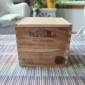 May include: A square wooden box with a light-colored lid and a darker wood body featuring finger-jointed corners. The front displays a black graphic of a villa with the text VILLA SAN LUCAS, a small rectangular stamp, and a circular brown wax seal.