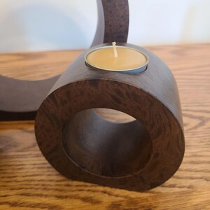 Op de afbeelding: Een donkerbruine houten kandelaar met een cirkelvormig ontwerp. De houder heeft een kleine metalen beker met een brandende, gele kaars. De houtnerf is zichtbaar. De kandelaar staat op een houten oppervlak.