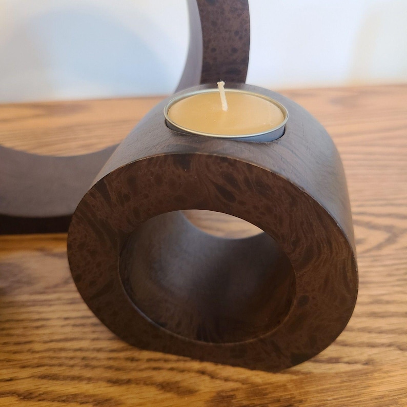 Op de afbeelding: Een donkerbruine houten kandelaar met een cirkelvormig ontwerp. De houder heeft een kleine metalen beker met een brandende, gele kaars. De houtnerf is zichtbaar. De kandelaar staat op een houten oppervlak.