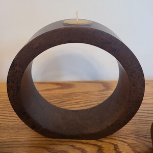 Op de afbeelding: Een donkerbruine, cirkelvormige houten kandelaar met een waxinelichtje. De kandelaar heeft een glad, gebogen ontwerp en staat op een houten oppervlak. Het waxinelichtje zit in een kleine, ronde metalen houder.