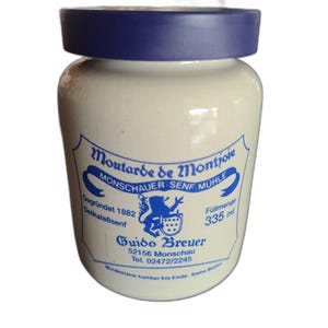 May include: A beige ceramic jar with a dark blue lid. The label features blue text reading Moutarde de Montjoie, Monschauer Senf Mühle, Gegründet 1882, Delikateßenf, Füllmenge 335 ml, Guido Breuer, 52156 Monschau, Tel. 02472/2245, and Mindestens haltbar bis Ende.