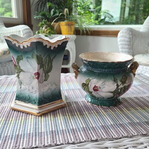 May include: A square-shaped ceramic vase with a scalloped rim and a rounded ceramic bowl with two side handles. Both pieces feature a gradient dark green to white finish, gold-painted trim, and a painted white hibiscus flower design with green leaves.