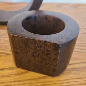 Op de afbeelding: Een bruine, geometrische kandelaar met een cirkelvormige opening. De houder heeft een vierkante basis en een gespikkeld patroon. De kandelaar is gemaakt van een donkerbruin materiaal en staat op een houten oppervlak.