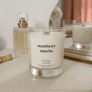 May include: A clear glass candle jar with a white label that reads "strawberry matcha". The label also includes the text "Hi Sunday Collective" and "coconut soy candle - wooden wick - 7oz - hand-poured in ny."