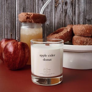 May include: A clear glass candle jar with a white label that reads "apple cider donut". The candle is surrounded by apples, a glass jar with a donut on top, and a white ceramic bowl with donuts.
