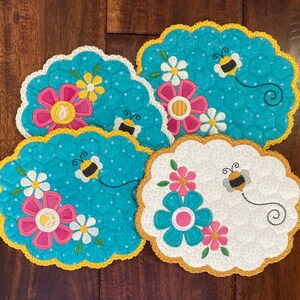 May include: Four quilted fabric coasters with a bee and flower design. The coasters are blue, white, and pink with yellow trim.