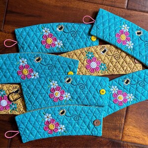 May include: Set of eight teal blue quilted coffee cup sleeves with embroidered flowers and bees. The sleeves have a honeycomb pattern and a pink loop handle.