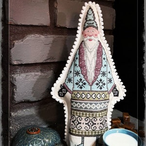 May include: A cross-stitch pattern of a blue and white Santa Claus with a white beard and a white hat. The Santa Claus is standing in front of a brick wall. The text "Selbu Santa" is at the top of the image and the text "By Cross Stitch Jo" is at the bottom of the image.