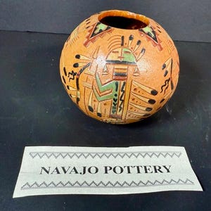 Puede incluir: Un jarrón redondo de cerámica Navajo de color naranja con una pequeña abertura. Está decorado con diseños geométricos y figurativos en negro, verde y marrón. El texto "NAVAJO POTTERY" está impreso en una etiqueta debajo.