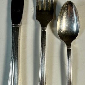 May include: A set of antique silverware, comprising a knife, fork, and spoon. The knife has a black blade, and all pieces feature ornate, detailed handles. The silverware is silver-coloured and likely made of metal.