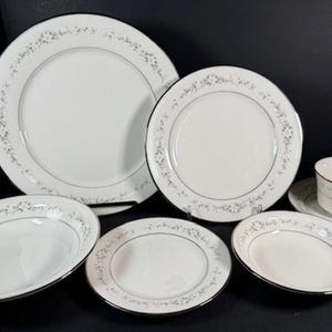 Op de afbeelding: Een set wit porselein serviesgoed met een zilveren bloemenpatroon. De set bevat een groot dinerbord, een saladebord, een soepkom, een brood- en boterbord, en een theekopje en schotel.