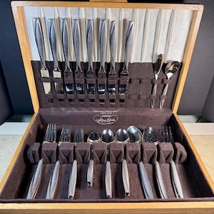 May include: A wooden silverware chest with a brown velvet interior, holding a complete set of stainless steel flatware. The set includes knives, forks, spoons, and serving utensils, all neatly arranged in their designated slots.
