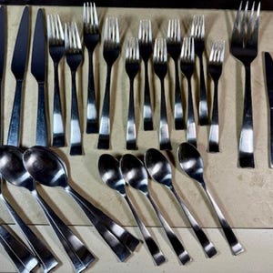 Könnte beinhalten: Eine Sammlung von Edelstahlbesteck, darunter Messer, Gabeln, Löffel und Teelöffel. Das Besteck hat ein schlankes, modernes Design mit poliertem Finish. Die Utensilien sind auf einer hellen Oberfläche angeordnet.