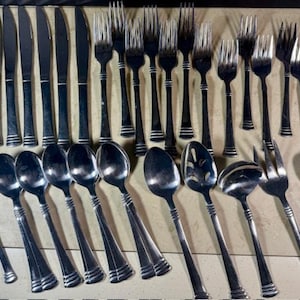 May include: A collection of stainless steel silverware, including knives, forks, and spoons, arranged on a light-colored surface. The silverware has a simple, elegant design with a shiny finish, suitable for formal dining.