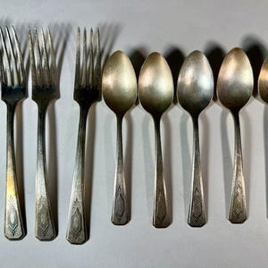 May include: A set of vintage cutlery, comprising forks and spoons. The forks have four prongs and ornate handles. The spoons feature oval bowls and long stems. The silverware is made of a silver-coloured metal and is arranged in a line.