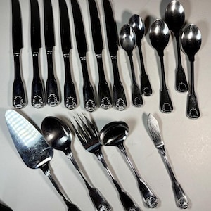 May include: A collection of vintage silverware, including knives, spoons, forks, and serving utensils. The silverware has a dark, possibly black, handle and a decorative shell design at the base. The spoons and forks have a shiny, reflective surface.