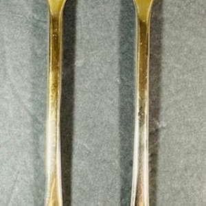 Pode incluir: Um conjunto de dois utensílios de servir dourados, um garfo e uma colher, com desenhos decorativos de conchas na parte inferior de cada cabo.