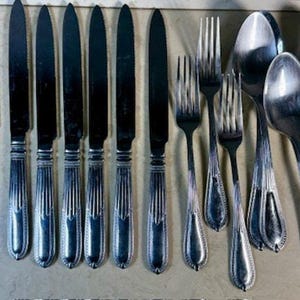 May include: A collection of vintage silverware, including knives, forks, and spoons. The stainless steel utensils have a decorative ribbed design on the handles. The knives have a dark, reflective blade. The arrangement suggests a formal dining setting.