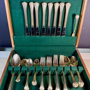 May include: A vintage cutlery set in a wooden case with a green velvet lining. The set includes multiple forks, spoons, and knives with ornate, gold-coloured handles. The case is open, displaying the arranged utensils.