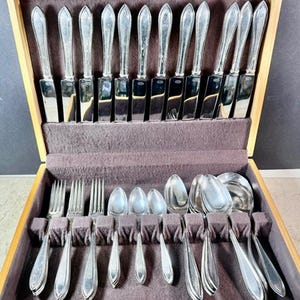 May include: A silver-toned flatware set arranged in a wooden storage case with a dark brown velvet lining. The set includes twelve knives with pointed blades, three forks, three small spoons, three medium spoons, and two large serving spoons.