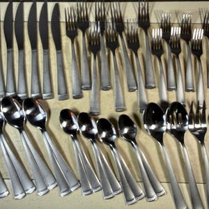 May include: A set of stainless steel cutlery, comprising knives, forks, and spoons, displayed on a light brown surface. The cutlery features a simple, elegant design with long, slender handles and polished heads.