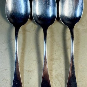 May include: Three vintage silver spoons with oval bowls and long, slender handles. The spoons are arranged vertically, showcasing their polished surfaces and classic design. The spoons are likely for serving or eating.