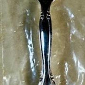Può includere: Un cucchiaio d'argento con un manico ornato e una grande ciotola ovale. Il cucchiaio è in un involucro di plastica trasparente. Il manico presenta un design floreale dettagliato. Questo cucchiaio è probabilmente per servire o mangiare.