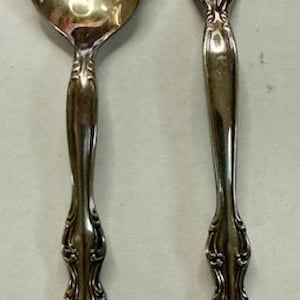Può includere: Set di cucchiaio e forchetta da portata argentati. Il cucchiaio ha una coppetta smerlata con intagli decorativi. La forchetta ha quattro rebbi. Entrambi i pezzi hanno manici ornati con motivi floreali.