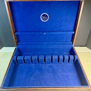 Può includere: Un portaposate in legno con interno in feltro blu. Il portaposate è aperto, rivelando scomparti per utensili. L'interno è blu scuro, in contrasto con l'esterno in legno. Un piccolo emblema circolare è visibile all'interno.