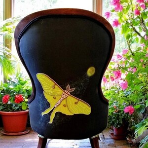 May include: Black upholstered chair with a large yellow moth design. The chair has a dark wooden frame and legs. The moth is detailed with yellow and brown accents. The chair is placed in a sunlit room with plants and flowers.