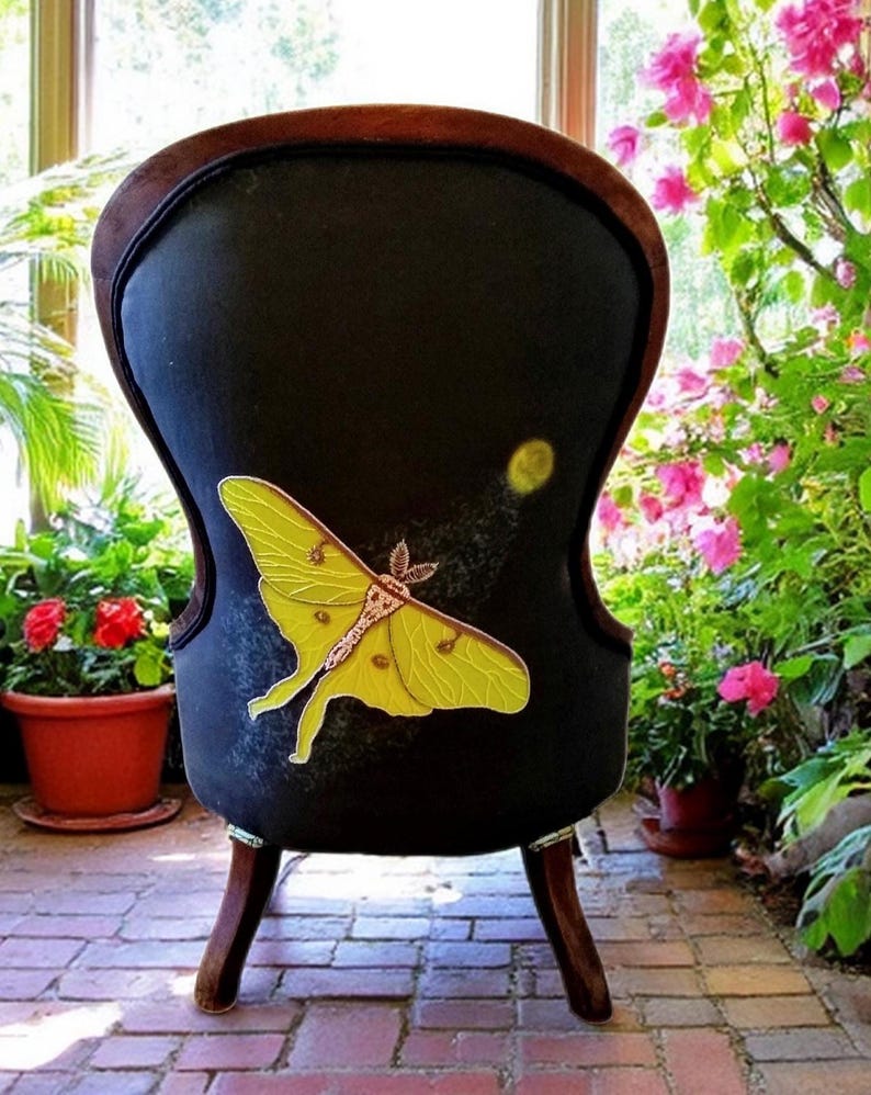 May include: Black upholstered chair with a large yellow moth design. The chair has a dark wooden frame and legs. The moth is detailed with yellow and brown accents. The chair is placed in a sunlit room with plants and flowers.