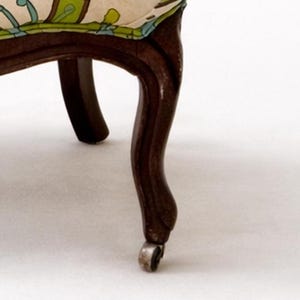 Pu&ograve; includere: Dettaglio di una sedia vintage con una gamba curva marrone scuro e una piccola rotella. Il sedile &egrave; rivestito in tessuto color crema con un motivo floreale verde, blu e giallo. Design classico.
