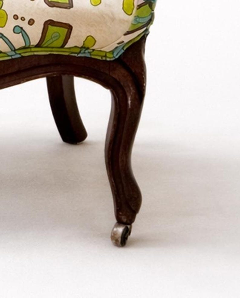Pu&ograve; includere: Dettaglio di una sedia vintage con una gamba curva marrone scuro e una piccola rotella. Il sedile &egrave; rivestito in tessuto color crema con un motivo floreale verde, blu e giallo. Design classico.