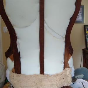 Pu&ograve; includere: Una sedia antica in legno in fase di restauro. La struttura marrone scuro sostiene un'imbottitura in schiuma bianca e una striscia di tessuto beige. Pezzi di schiuma e ritagli di tessuto sparsi.