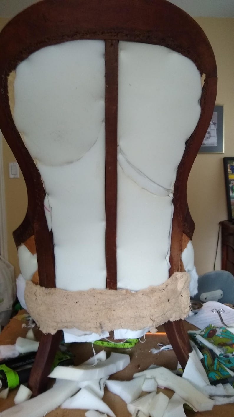 Pu&ograve; includere: Una sedia antica in legno in fase di restauro. La struttura marrone scuro sostiene un'imbottitura in schiuma bianca e una striscia di tessuto beige. Pezzi di schiuma e ritagli di tessuto sparsi.