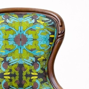 Pu&ograve; includere: Primo piano dello schienale di una sedia vintage con una cornice in legno intagliato. Lo schienale della sedia presenta un tessuto colorato a fantasia nei toni del verde, blu, giallo e marrone. La cornice in legno ha una finitura marrone scuro.