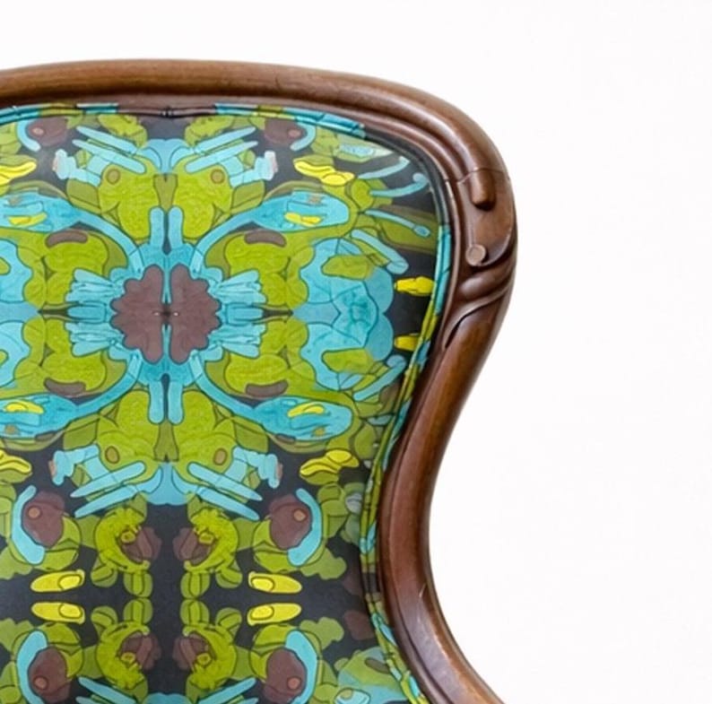 Pu&ograve; includere: Primo piano dello schienale di una sedia vintage con una cornice in legno intagliato. Lo schienale della sedia presenta un tessuto colorato a fantasia nei toni del verde, blu, giallo e marrone. La cornice in legno ha una finitura marrone scuro.