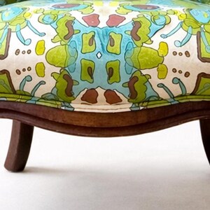 Pu&ograve; includere: Primo piano di una sedia vintage con seduta fantasia. Il tessuto presenta un motivo ripetuto nei toni del verde, blu e marrone su sfondo crema. La sedia ha gambe in legno marrone scuro.