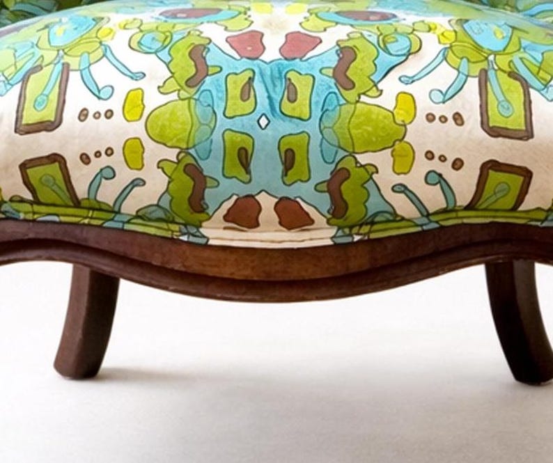 Pu&ograve; includere: Primo piano di una sedia vintage con seduta fantasia. Il tessuto presenta un motivo ripetuto nei toni del verde, blu e marrone su sfondo crema. La sedia ha gambe in legno marrone scuro.