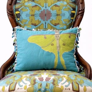 Pu&ograve; includere: Un cuscino turchese decorativo con un grande disegno di falena gialla, rifinito con bordi bianchi e nappe blu. Il cuscino &egrave; posto su una sedia fantasia con una cornice in legno, creando un'estetica vintage.