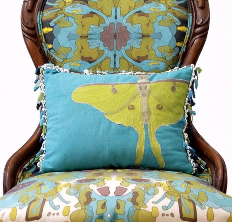 Pu&ograve; includere: Un cuscino turchese decorativo con un grande disegno di falena gialla, rifinito con bordi bianchi e nappe blu. Il cuscino &egrave; posto su una sedia fantasia con una cornice in legno, creando un'estetica vintage.
