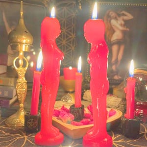 Puede incluir: Dos velas rojas con forma humana se encuentran frente a frente, cada una con una llama encendida. Están rodeadas de velas de pilar rojas más pequeñas en soportes negros con símbolos de pentáculo, un soporte de metal dorado y pétalos rosas.