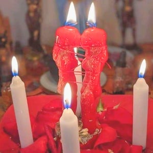 May include: Two red figure-shaped candles stand side-by-side, connected by thin red wax strands, with lit wicks. They are surrounded by three white pillar candles and scattered red rose petals, all illuminated by the flickering flames.