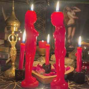 Puede incluir: Dos velas rojas con forma de figura humana, con mechas encendidas, destacan, flanqueadas por velas rojas más pequeñas en soportes negros. El conjunto está dispuesto sobre una mesa con un quemador de incienso dorado y una obra de arte enmarcada al fondo.