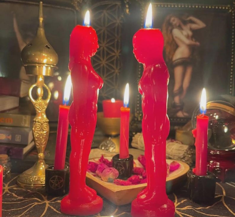 Pu&ograve; includere: Due candele rosse a forma di figura umana con stoppini accesi sono in evidenza, affiancate da candele rosse pi&ugrave; piccole in supporti neri. Le candele sono su un tavolo con altri oggetti spirituali, tra cui un bruciatore d'incenso dorato.